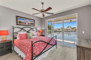 A bedroom with a bed, a ceiling fan, a nightstand with a lamp, and a view of a body of water through large glass doors.
