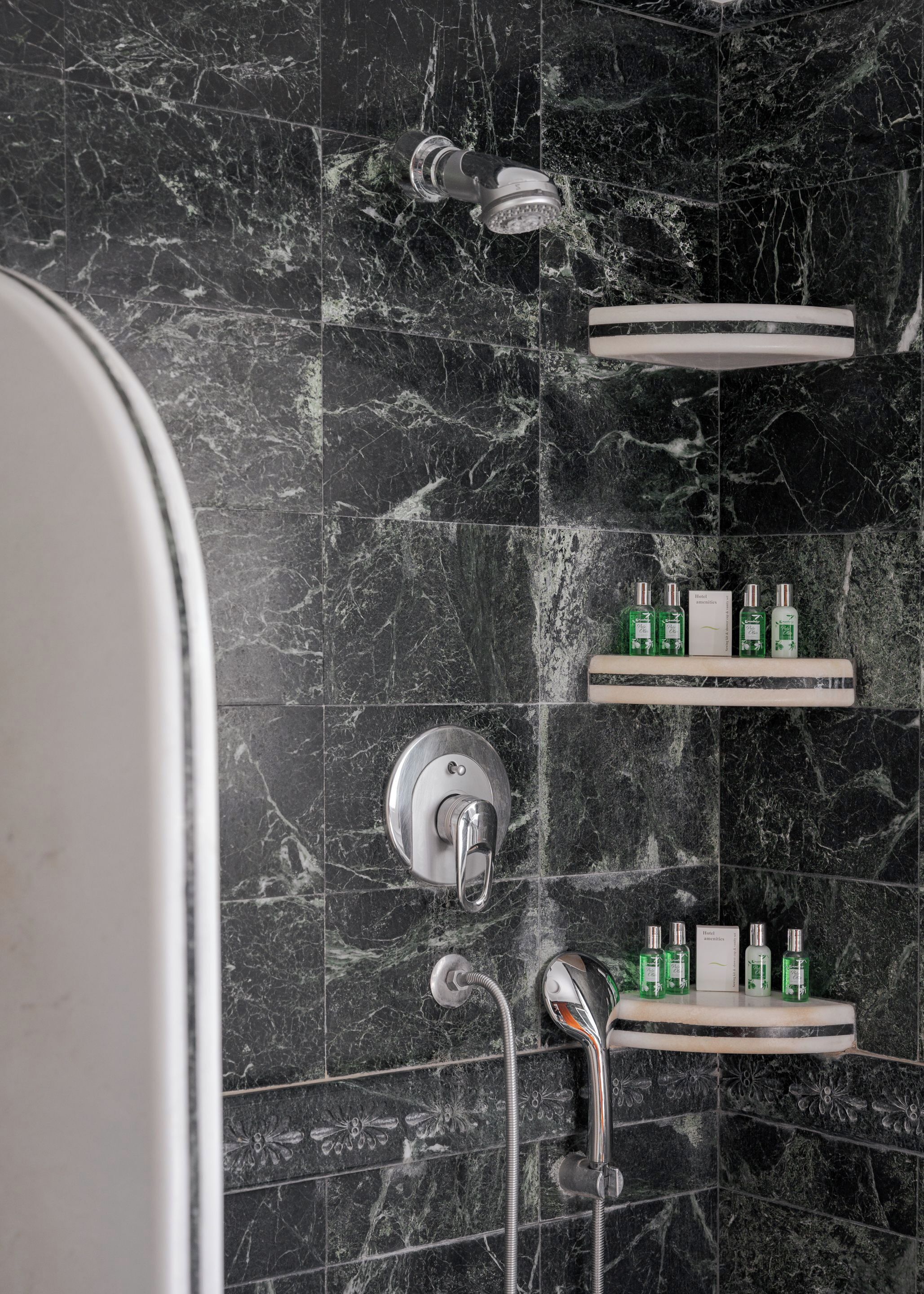 Une salle de bain avec un mur en marbre noir, une douche équipée d’un pommeau de douche à main et d’un robinet mural. On y trouve également des étagères contenant des articles de toilette.