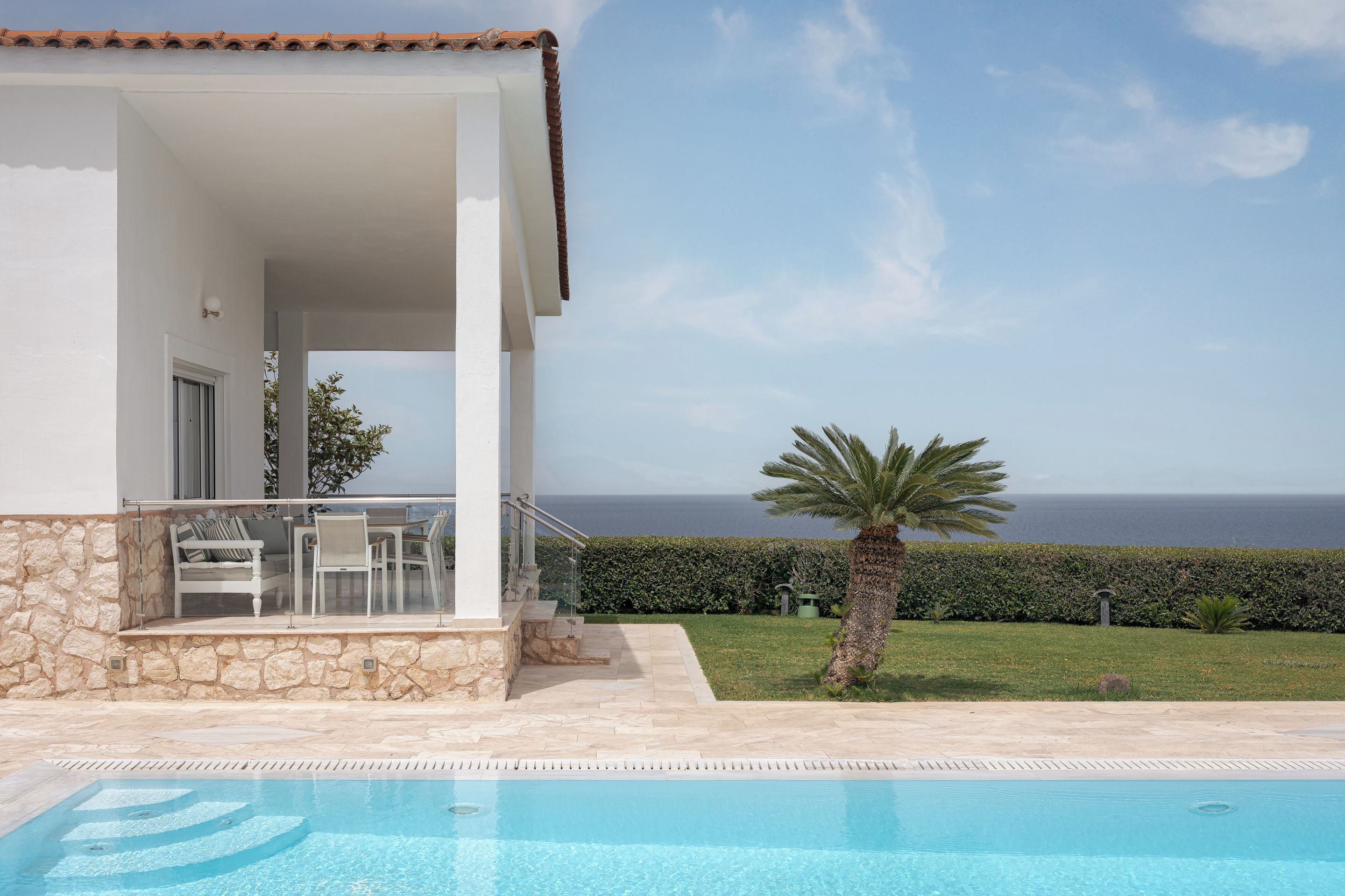 A poolside area with a view of the sea, a stone wall, and a palm tree.