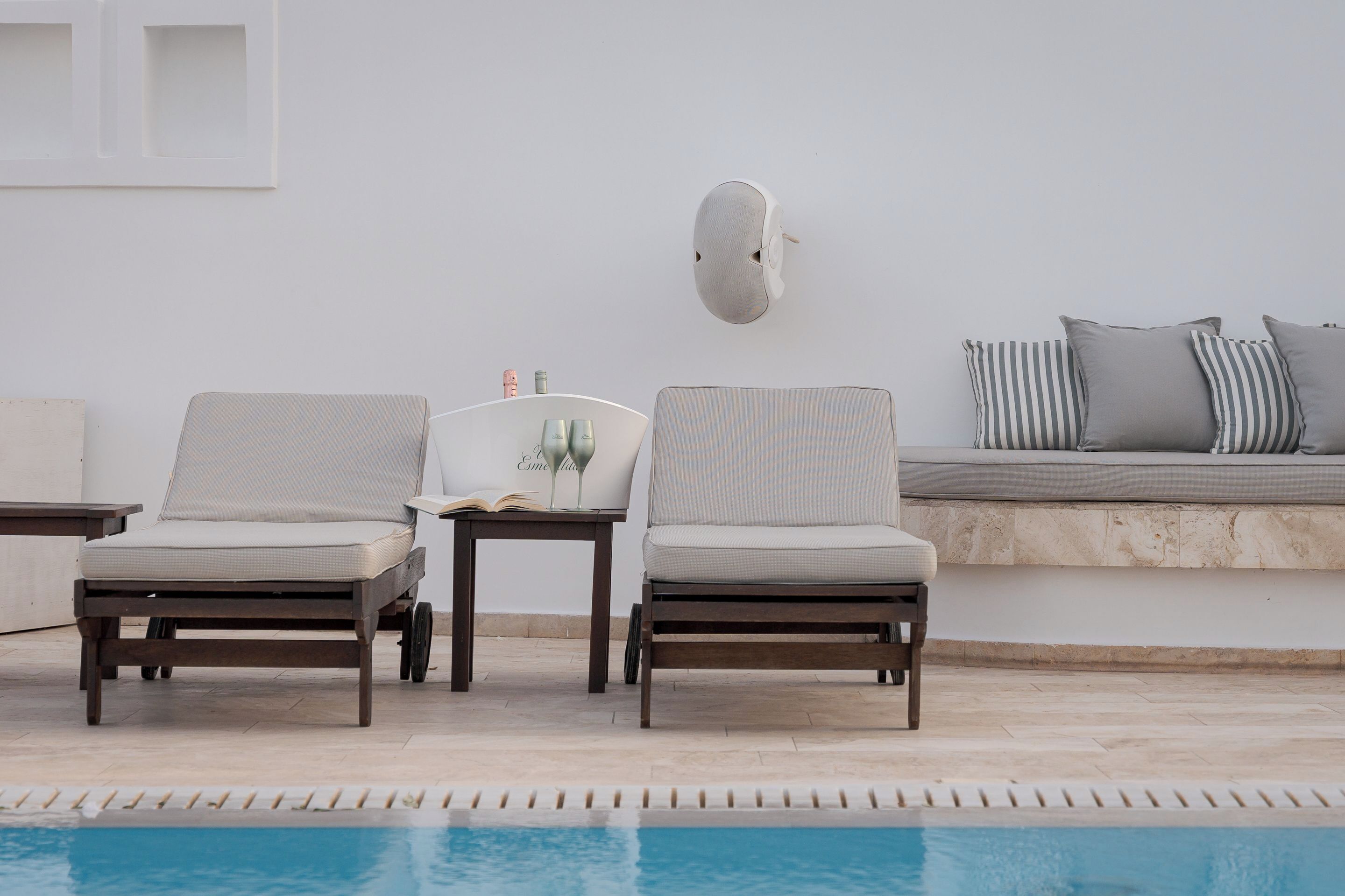 A poolside area with two lounge chairs, a small table with a book and glasses, and a wall-mounted light fixture.