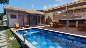 Une piscine avec un bord carrelé, un patio couvert comprenant une table et des chaises, et un petit bâtiment doté d’une porte et de fenêtres.