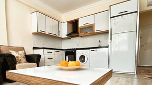 Eine moderne Küche mit weißen Schränken, einer eingebauten Mikrowelle und einem Kühlschrank. Ein Holztisch mit einer Fruchtschale steht daneben.