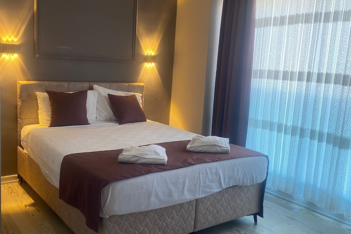 Habitació d'hotel amb un llit gran, una televisió plana penjada a la paret sobre el llit i una finestra amb cortines translúcides.