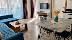 Ein modernes Wohnzimmer mit einer Couch, Couchtisch, Esstisch und Stühlen. Ein Fernseher ist an der Wand montiert und eine Tür mit Griff ist zu sehen.