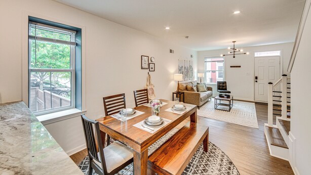 A spacious dining area with a wooden table, chairs, and a bench. There is a window with blinds, a sofa, and a staircase in the background. - Chic Lancaster Vacation Rental, Walkable Location! (Lancaster)