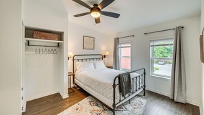 A bedroom with a bed, a ceiling fan, a window with curtains, a bedside table, and a shelf with hanging items. - Chic Lancaster Vacation Rental, Walkable Location! (Lancaster)