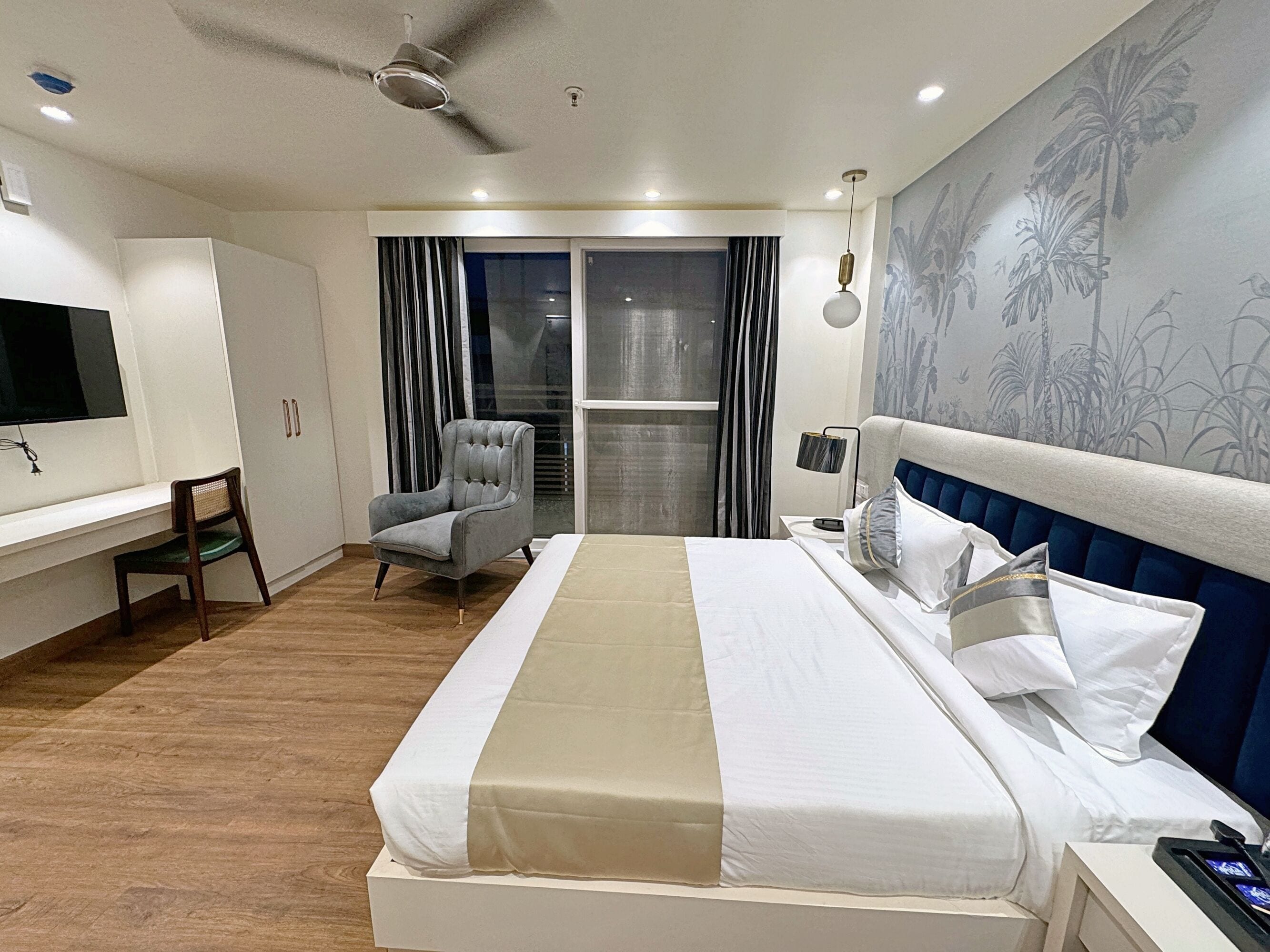 A modern hotel room with a large bed, a desk, a chair, and a view of the outdoors through a sliding door.