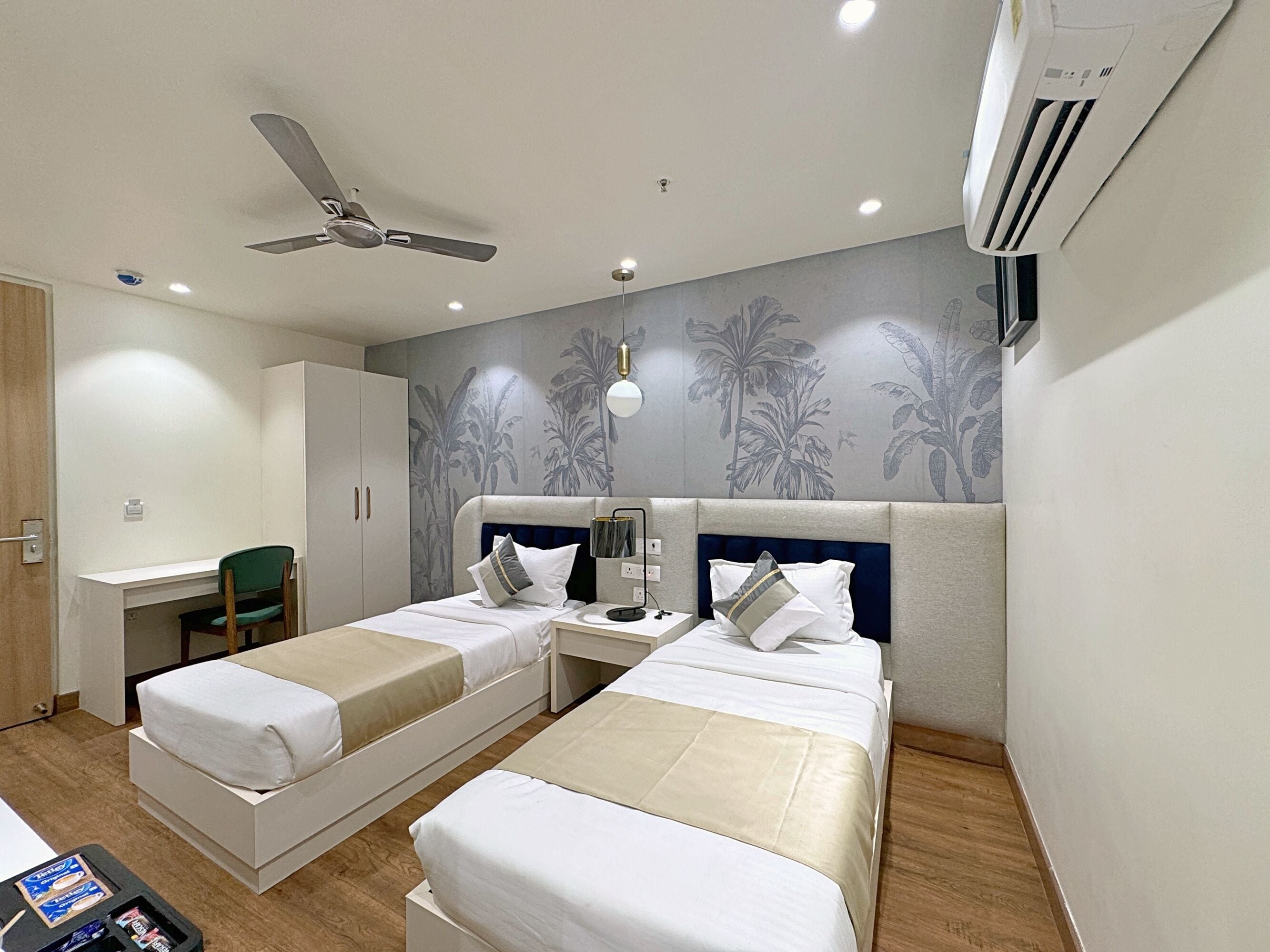 A hotel room with two single beds, a desk, a chair, a ceiling fan, and an air conditioning unit.