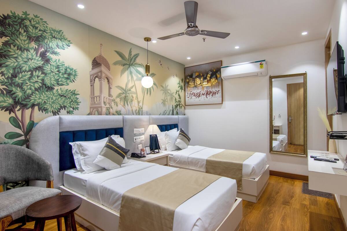 Ein Hotelzimmer mit zwei Betten, einem Deckenventilator, einem Wandbild von Bäumen und einem Tempel sowie einem Fernseher.