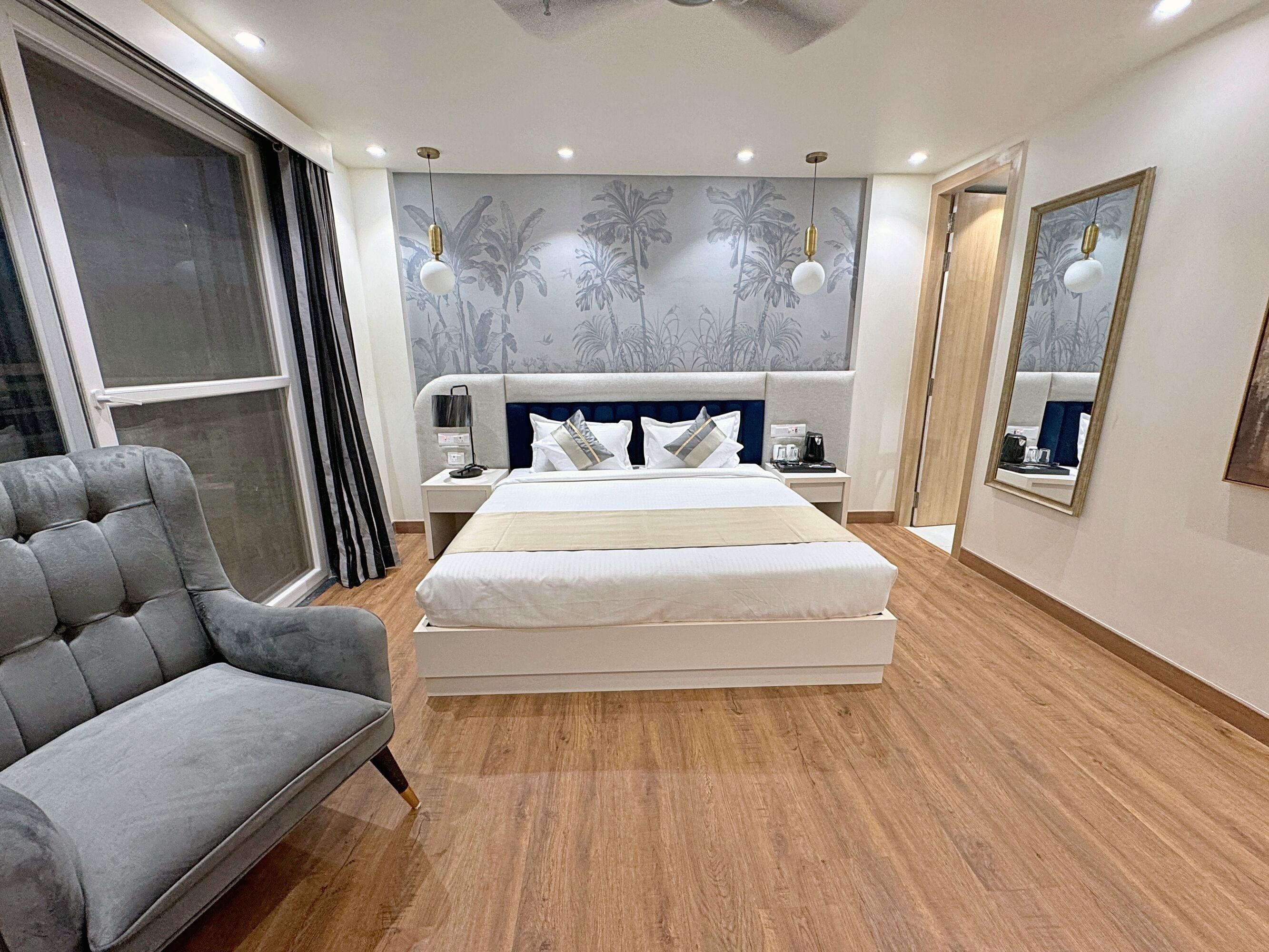 A modern hotel room with a large bed, a grey armchair, a nightstand with a lamp, and a wall with a tropical mural.
