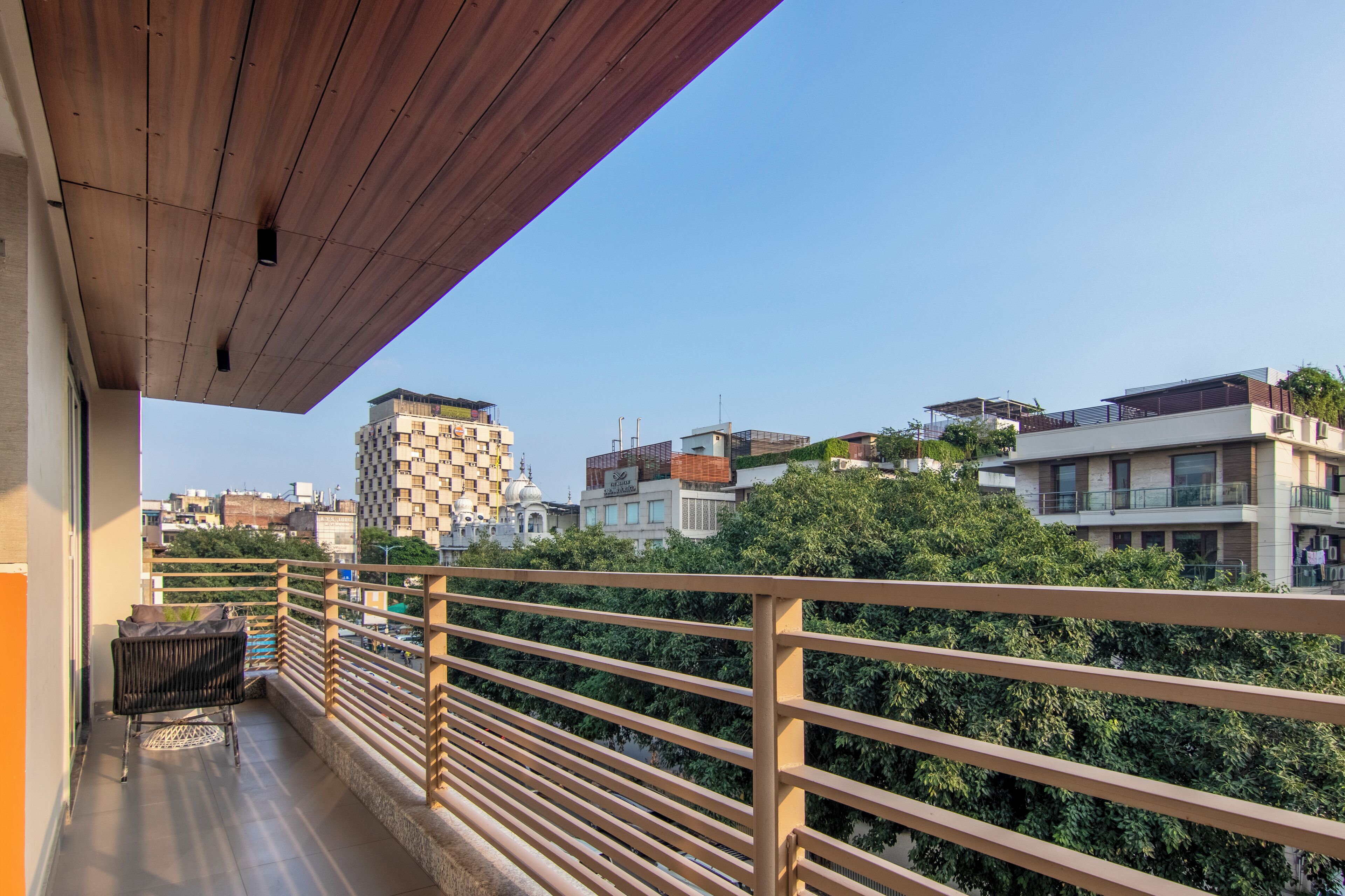 A balcony with a wooden railing, a wicker chair, and a view of modern buildings and greenery.