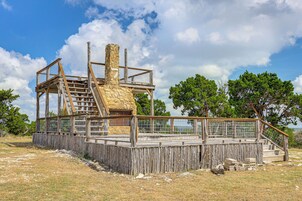 Una construcción rústica de madera con chimenea de piedra, escaleras de madera y una zona delimitada por una valla.