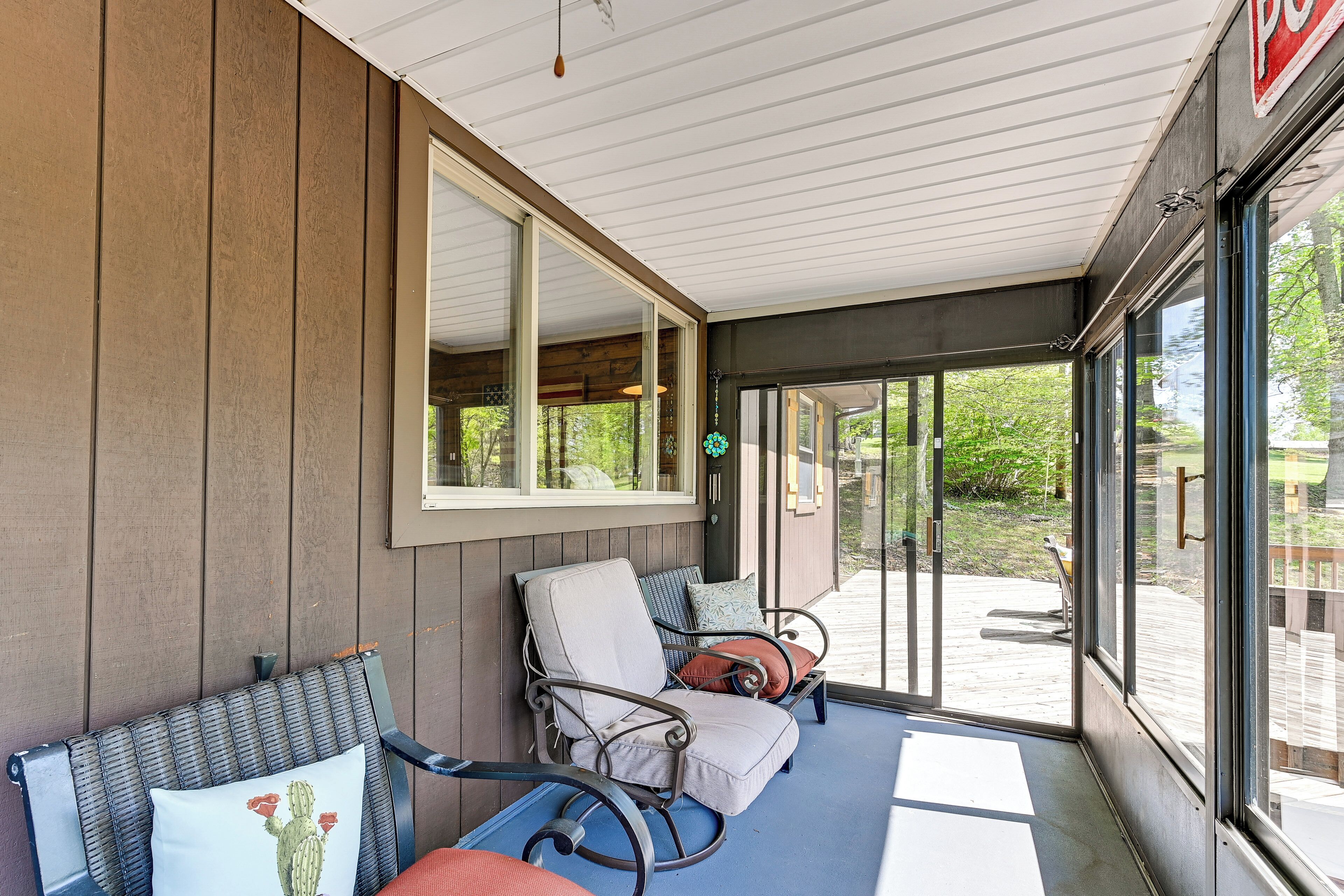 A covered outdoor seating area with two rocking chairs and a bench, large windows, and a view of a wooden deck and greenery.