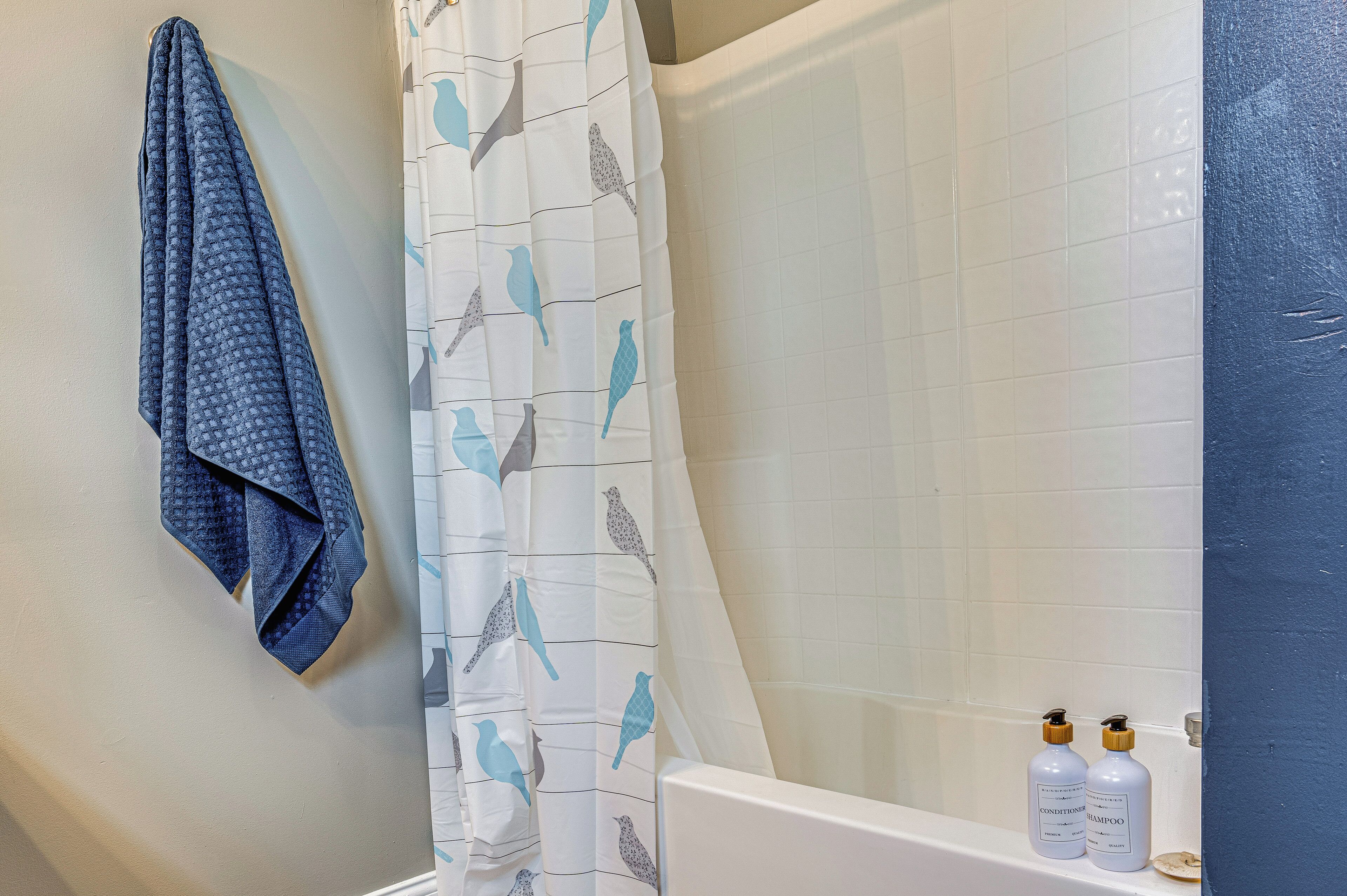 A bathroom with a shower curtain featuring bird patterns, a towel hanging on the wall, and two bottles of shampoo and conditioner on the bathtub.