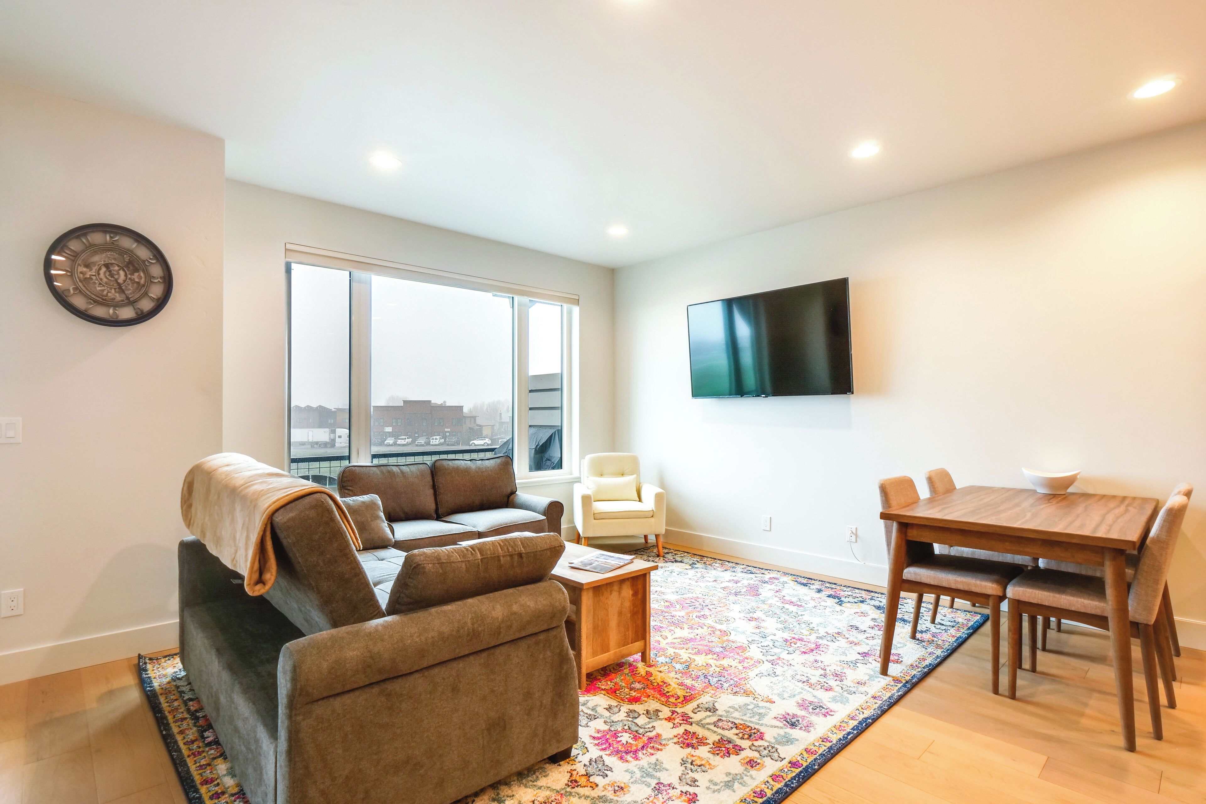 A modern living room with a sofa, coffee table, and a dining table with chairs. A flat-screen TV is mounted on the wall.
