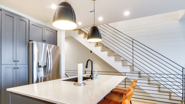 Una cocina moderna con un refrigerador de acero inoxidable, una encimera blanca y un lavamanos empotrado. Dos lámparas colgantes iluminan la encimera y una escalera con barandilla metálica conduce a un nivel superior.