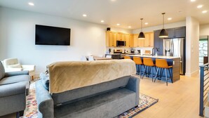 Una sala moderna con un sofá gris, un televisor de pantalla plana en la pared y una zona de cocina con armarios de madera, un refrigerador y taburetes de bar.