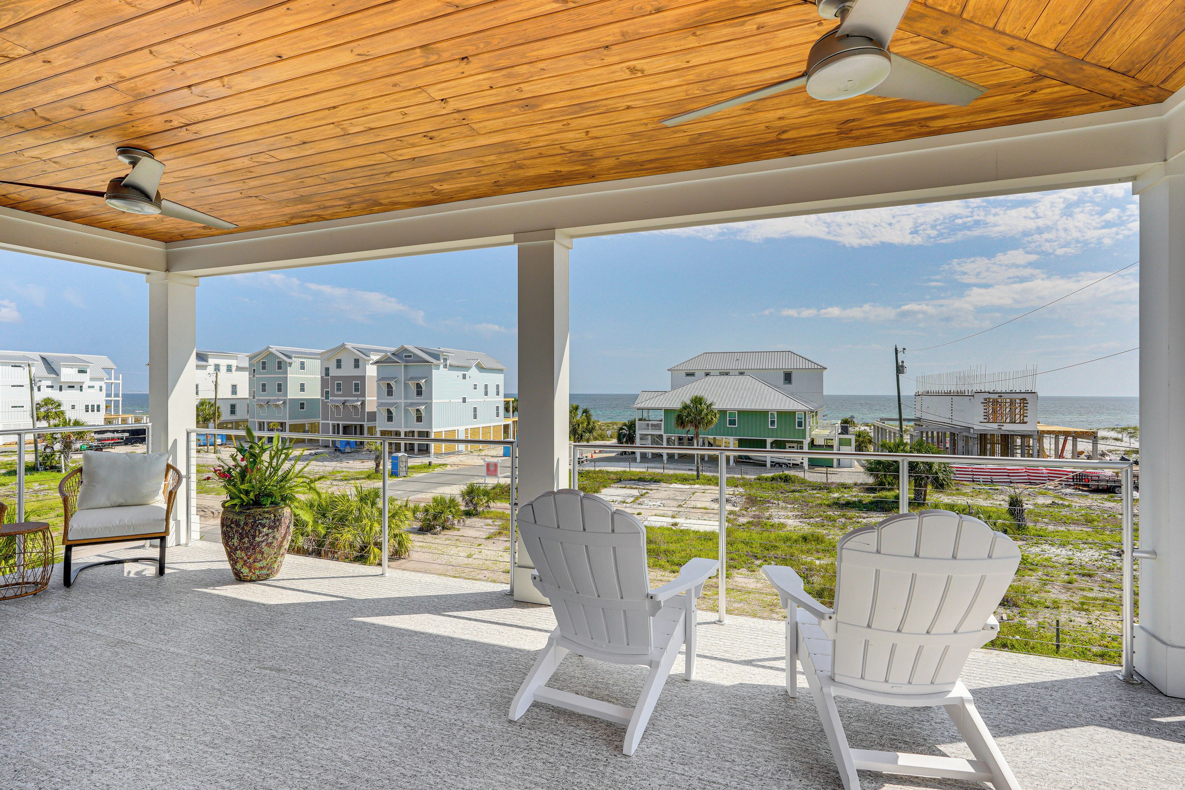 Ein überdachter Außenbereich mit zwei weißen Adirondack-Stühlen, einem Blumentopf und Blick auf Strand und Gebäude im Hintergrund.