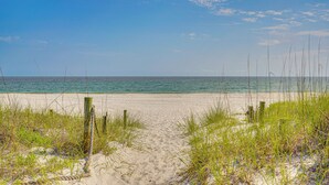 Un sentier de sable qui longe la plage et mène à l’océan, bordé de gazon et de poteaux en bois.