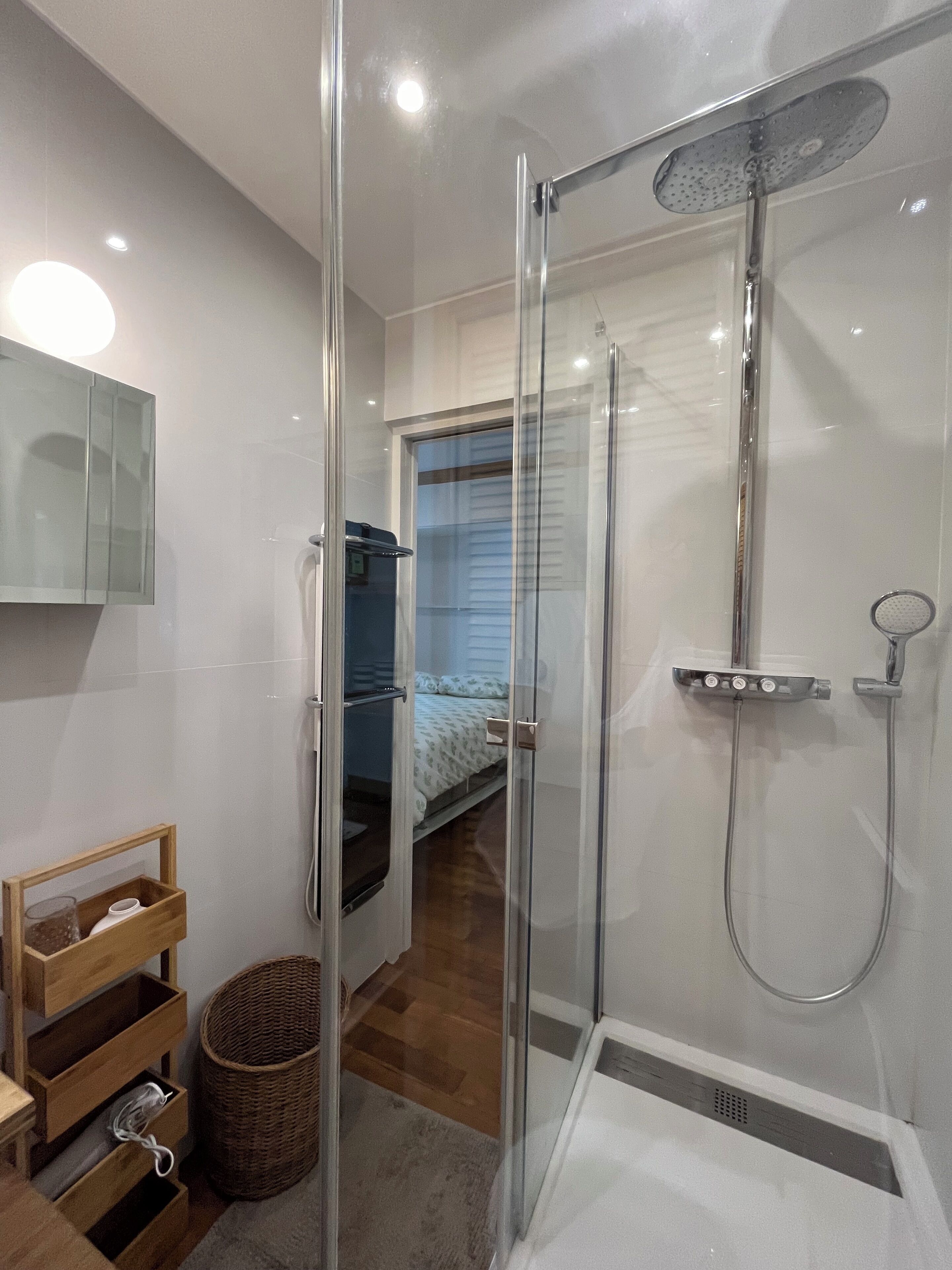 A modern bathroom with a glass-enclosed shower, a wooden vanity, and a visible bedroom in the background.