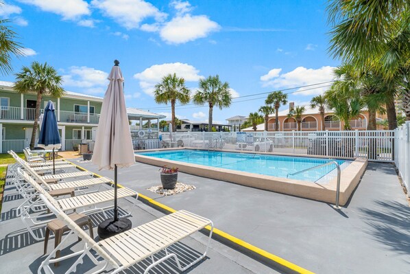 A pool area with lounge chairs and umbrellas, surrounded by a fence and palm trees.