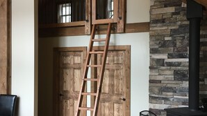 Une chambre à coucher aménagée dans un espace mansardé, avec une échelle en bois, une cheminée en pierre et un coin salon confortable comprenant un canapé et un fauteuil.