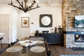 A dining area with a wooden table, chairs, and a decorative centerpiece. There is a stone fireplace with a fire, a large mirror, and a framed artwork on the wall. - 12 Wildwood 2 Bedroom Condo by Moonlight Basin Lodging (Big Sky)