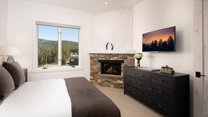Un dormitorio con una cama grande, chimenea, cómoda, televisor y ventana con vista a árboles y una casa.