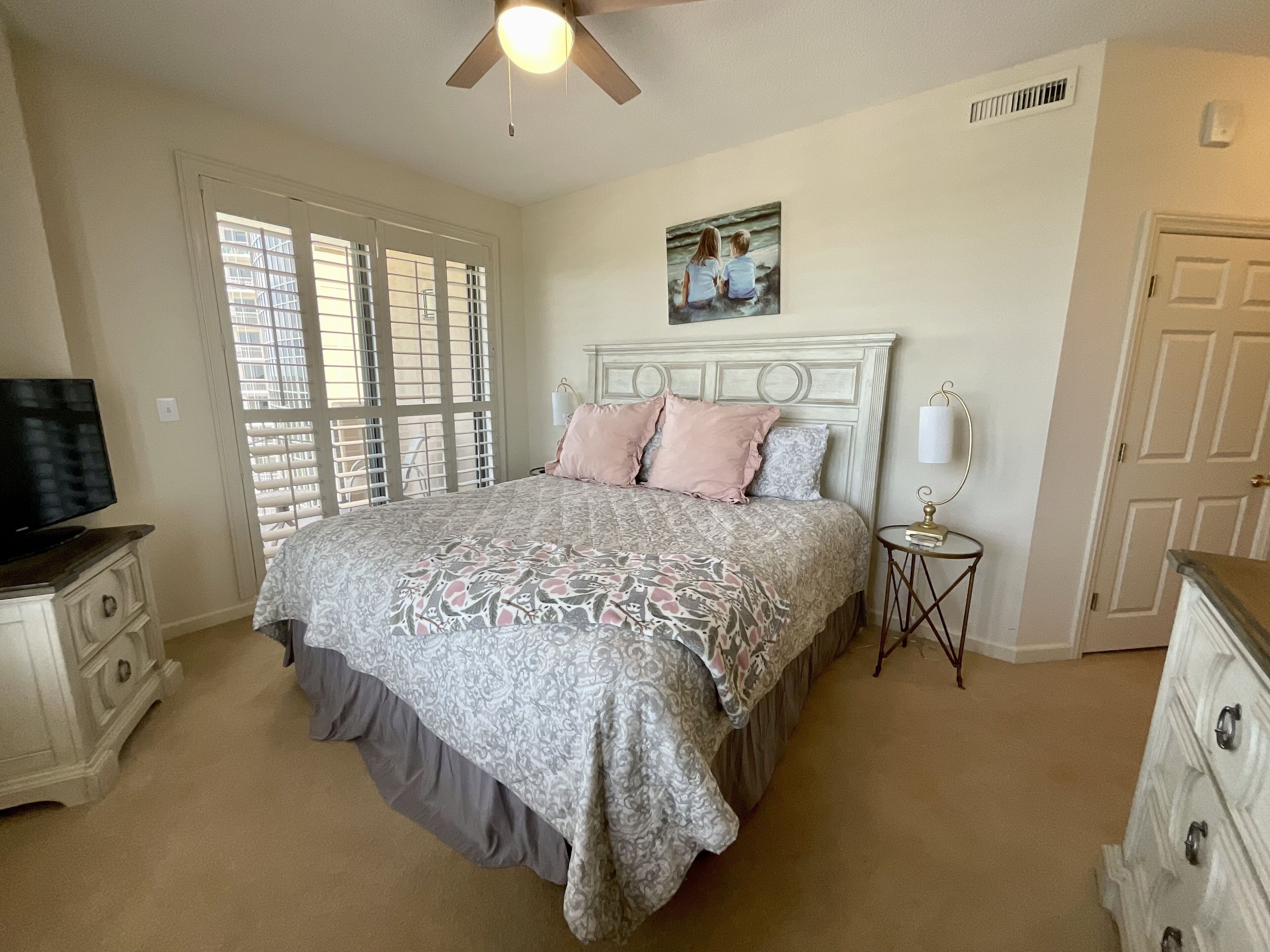 A bedroom with a large bed, a ceiling fan, a painting above the bed, a television on a dresser, and a door.