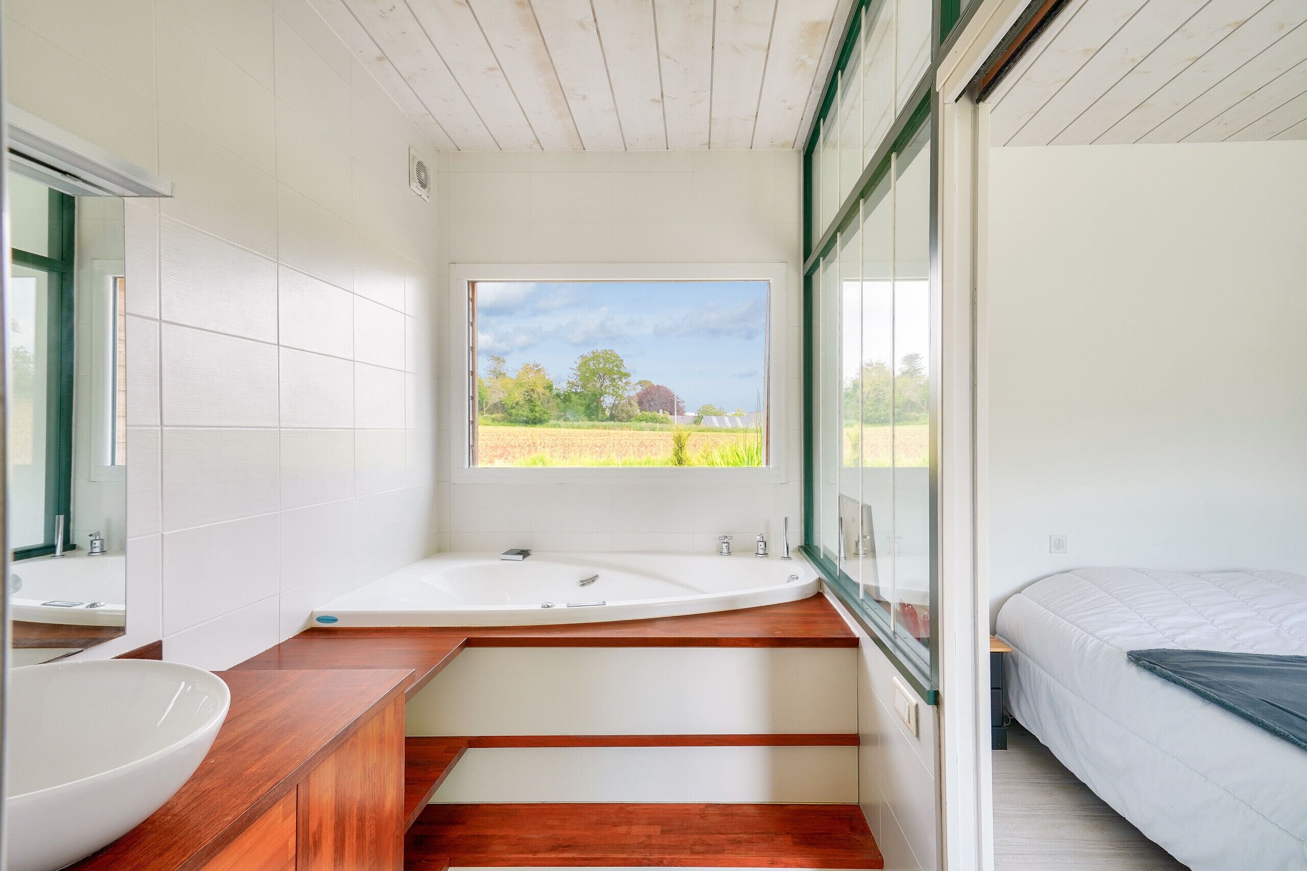 A modern bathroom with a large bathtub, a wooden vanity, and a large window offering a view of a field.