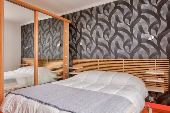 A bedroom with a mirrored wardrobe, a bed with a white comforter, and a patterned wallpaper.