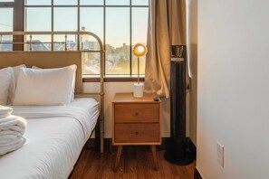 Une chambre d’hôtel avec un lit, une table de chevet avec une lampe, un radiateur et une fenêtre donnant sur la ville.