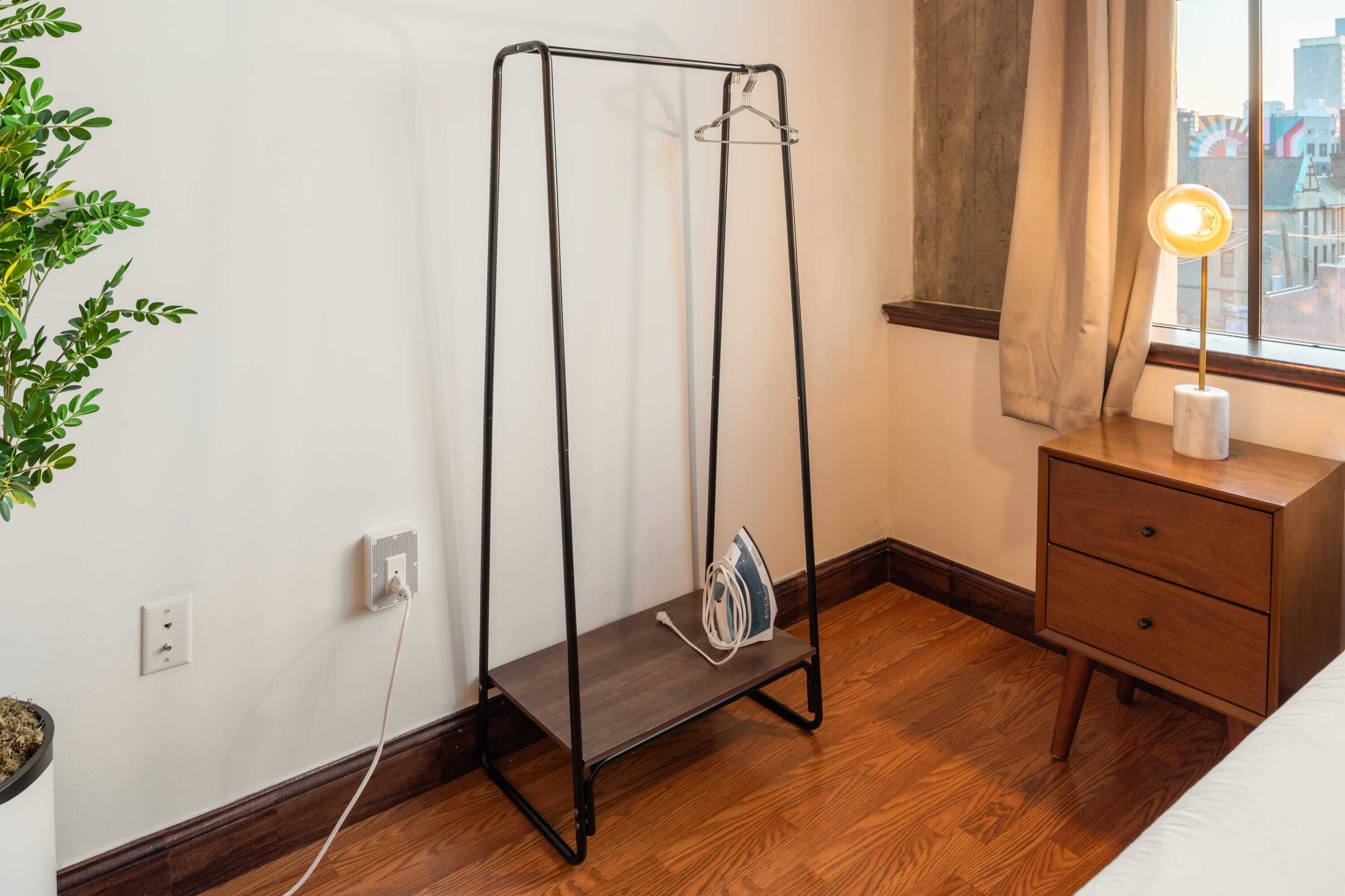 Una habitación con un tendedero de metal, una mesita de noche con lámpara, una ventana con cortinas y una planta.