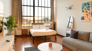 A modern hotel room with a large bed, a sofa, a small table with a plant, a window with curtains, and a city view. - Sosuite | Studio w W/D, Gym, On-Site Restaurant (Philadelphia)