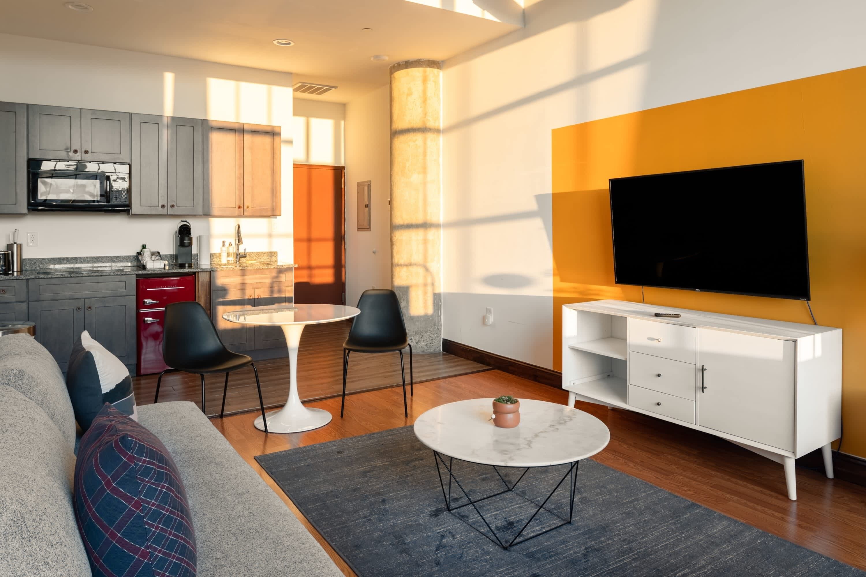 A modern living room with a sofa, two chairs, a coffee table, and a TV. There is a kitchen area with cabinets and appliances.