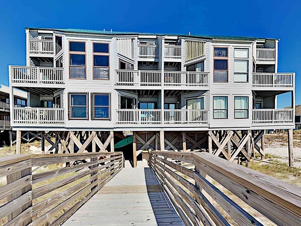 A multi-story wooden building with balconies, a wooden walkway leading to it, and a clear blue sky in the background.