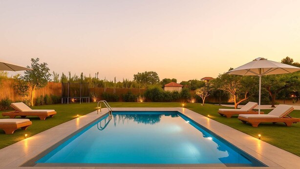 A rectangular swimming pool with a clear blue water surface, surrounded by a paved deck. There are lounge chairs with white cushions and wooden frames on both sides of the pool.