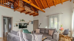 A spacious living room with a high wooden ceiling, a grey sofa set, a television, and a dining area with a table and chairs.