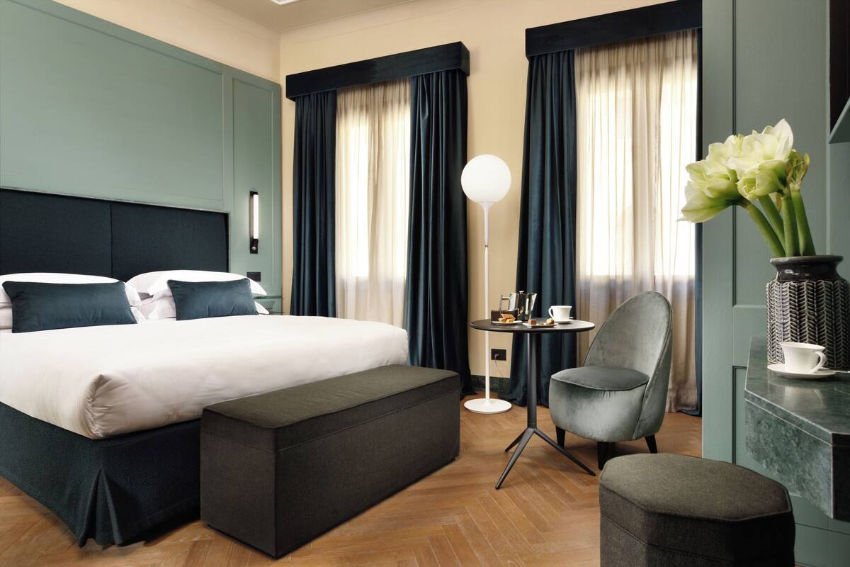 A hotel room with a large bed, a small table with a lamp, a chair, and a vase with flowers.