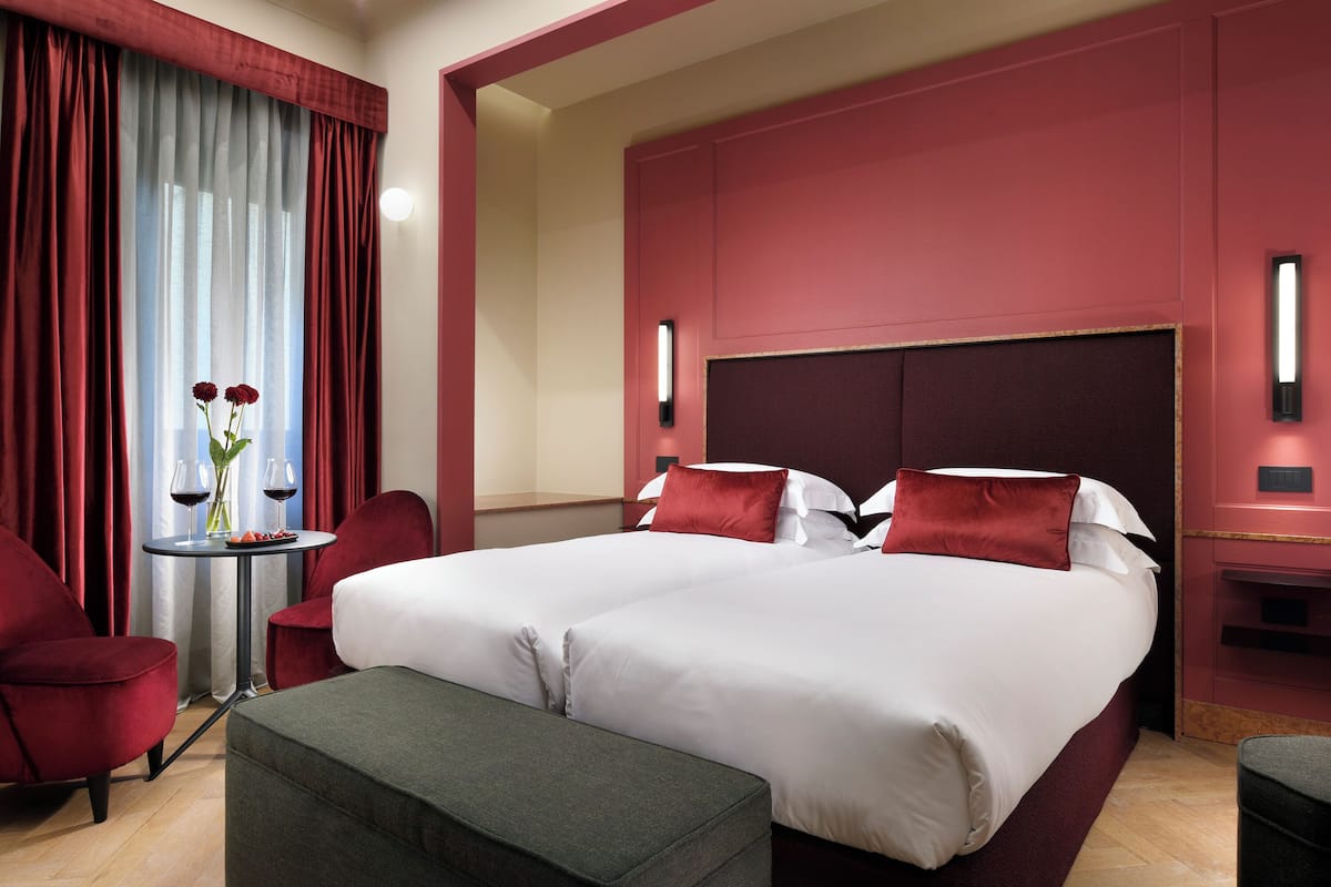 A hotel room with a large bed, two red chairs, a small table with a vase and wine glasses, and a bench.