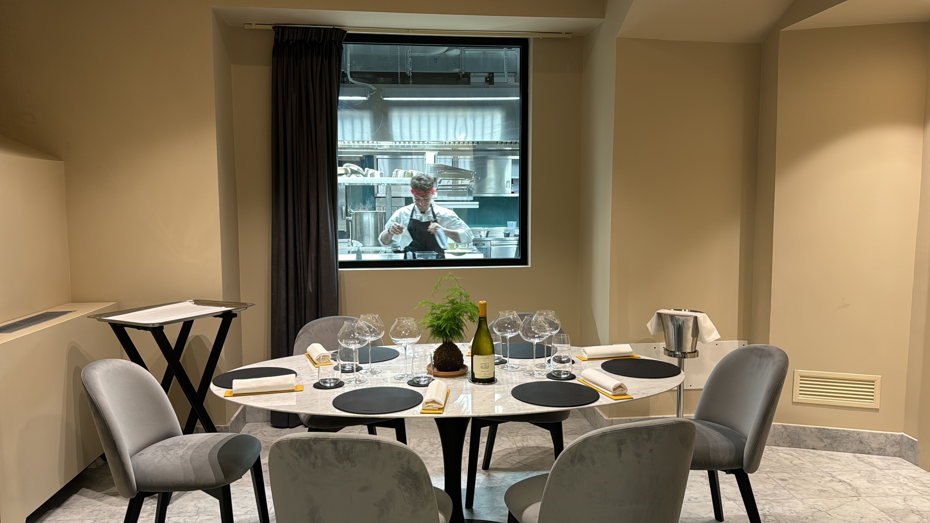Uma sala de conferências com uma mesa redonda, quatro cadeiras, uma tela de projetor e uma janela com vista para uma cozinha.