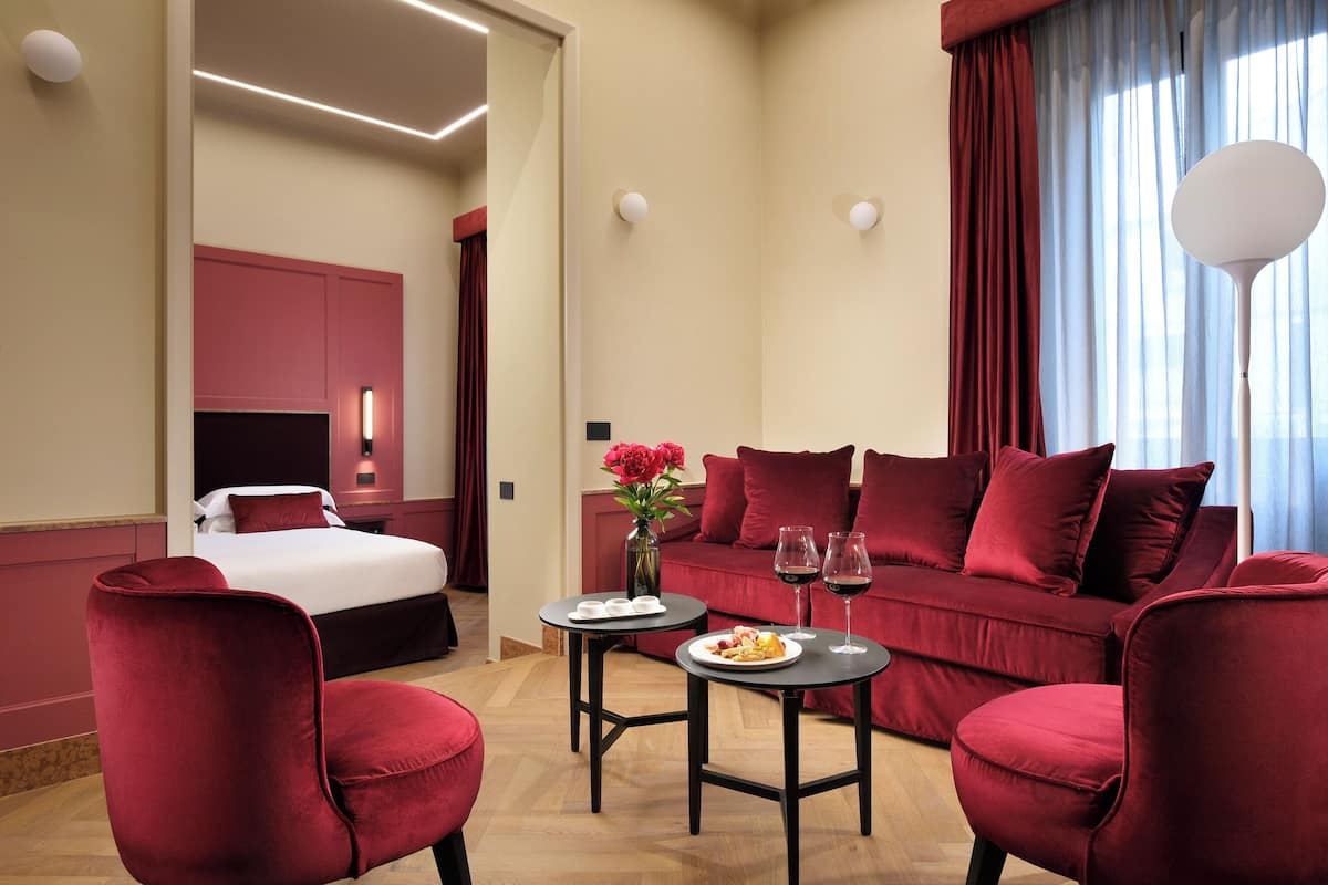 A hotel room with a bed, a red sofa, two armchairs, a small table with a plate of food and two wine glasses, and a vase with flowers.
