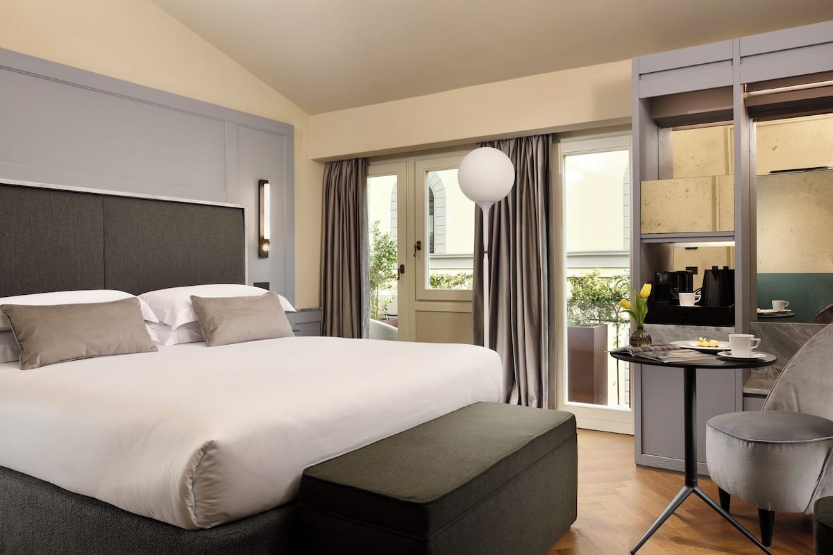 A modern hotel room with a large bed, a small table with a cup and saucer, a chair, and a balcony with curtains.
