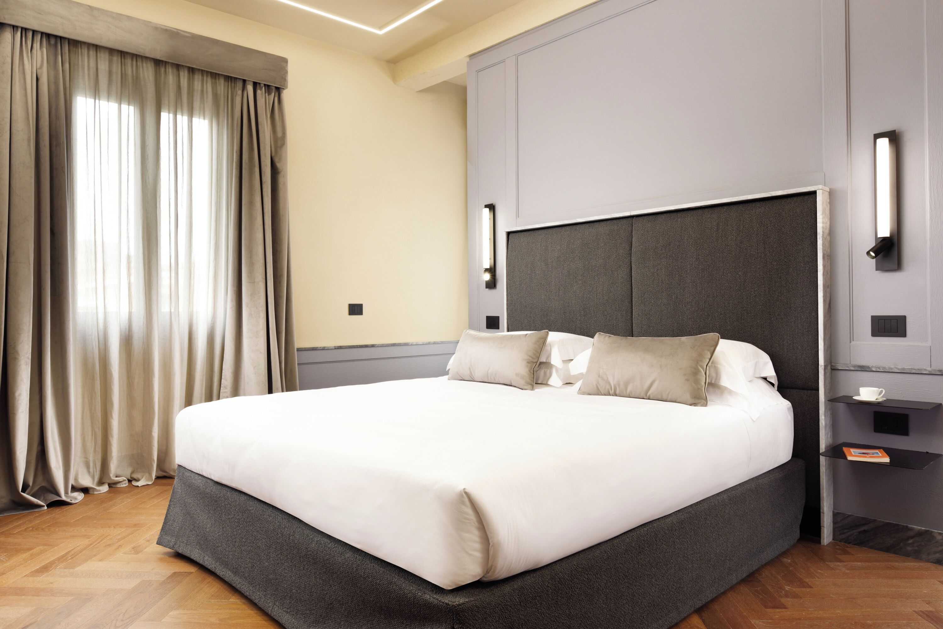 Een moderne hotelkamer met een groot bed, een grijs hoofdeinde en een nachtkastje met een lamp.