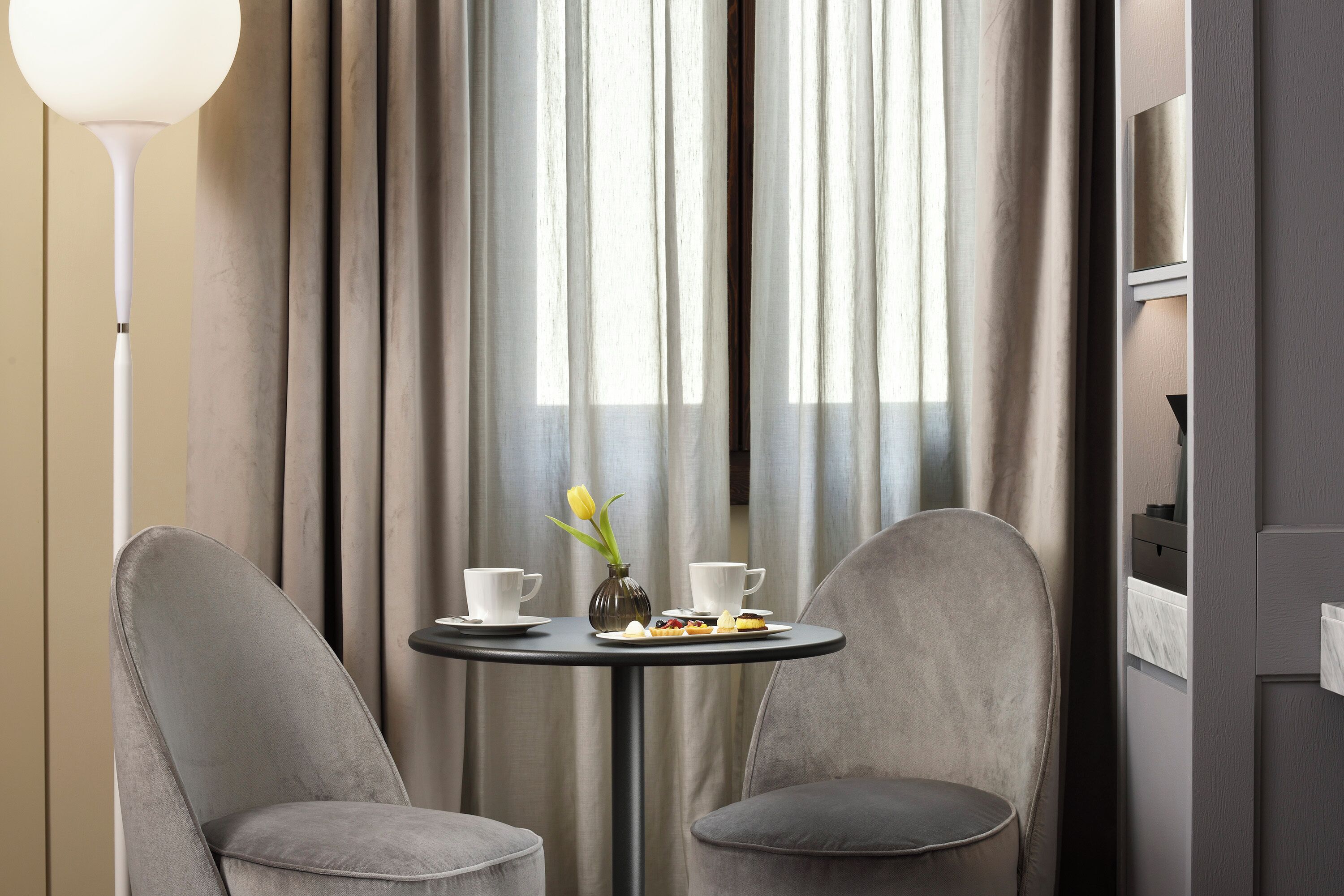 A modern room with two grey upholstered chairs, a small round table with a vase of yellow tulips, two white cups, and a plate of food.