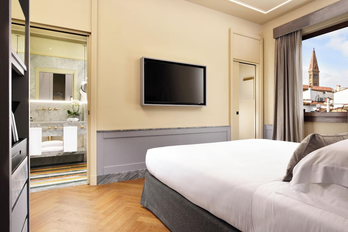 A hotel room with a large bed, a flat-screen TV mounted on the wall, a bathroom with a mirror and sink, and a view of a building outside.