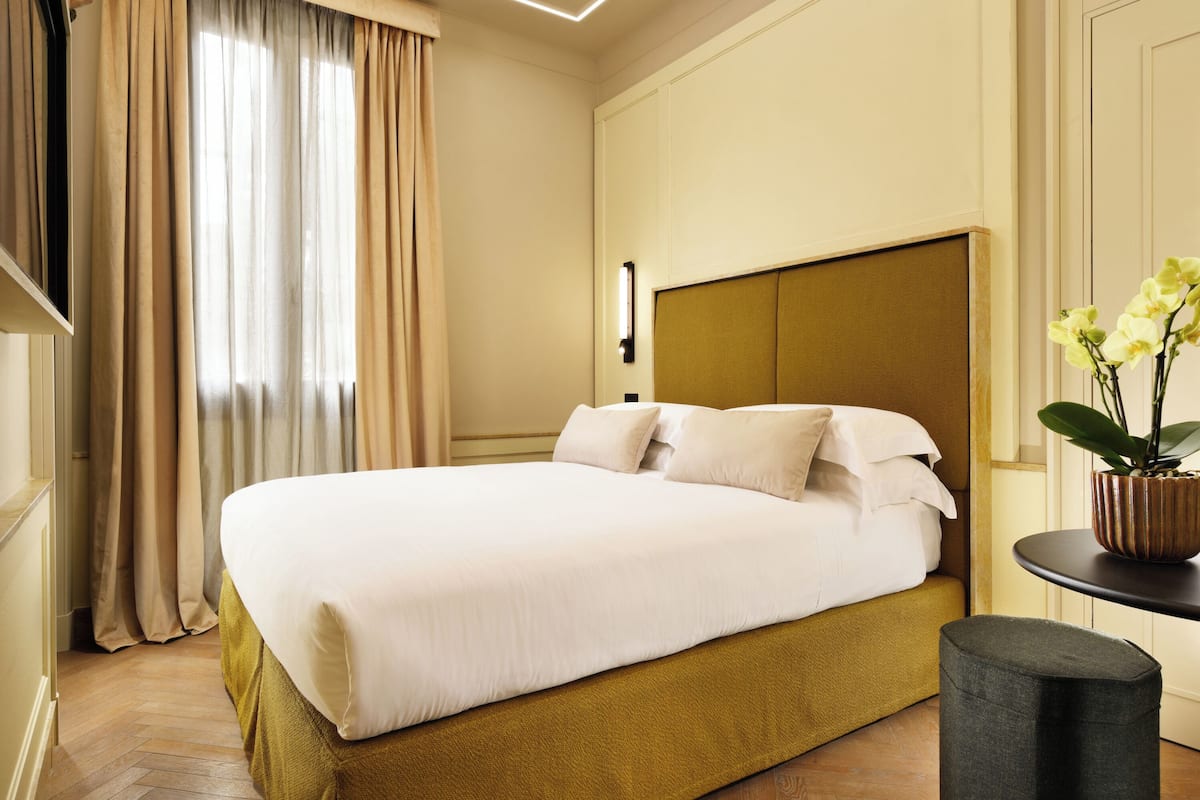 A neatly made bed with a headboard, a potted plant on a side table, and a window with curtains.