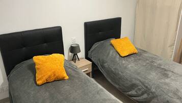 Une chambre double avec deux lits, chacun étant recouvert d’une couverture grise et agrémenté d’un oreiller jaune. Une petite table de chevet avec une lampe se trouve entre les deux lits.