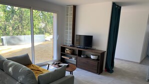 Una sala de estar moderna con un sofá gris, una mesa de centro de madera, un televisor sobre un mueble de madera y un ventanal corredero que da a un espacio al aire libre.