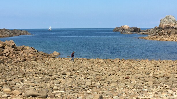 En stenet strand med en person, en sejlbåd i det fjerne og store sten.